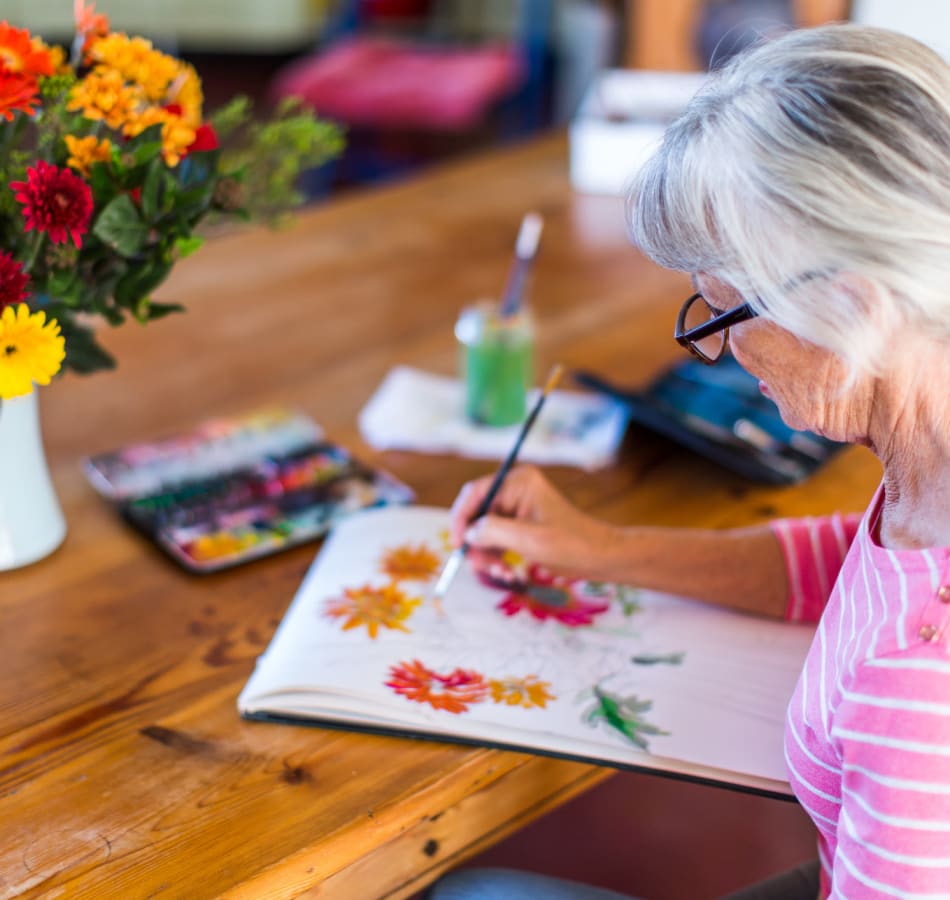 A senior sits at a picnic table in an assisted living community painting in the afternoon sun